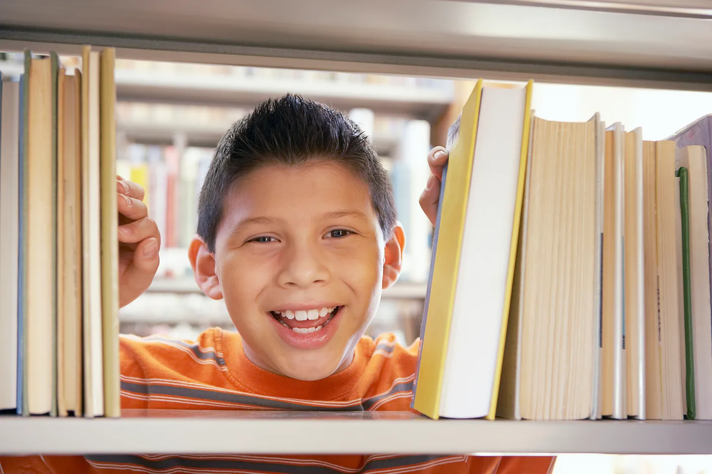 A smiling boy peeking through books on a library shelf