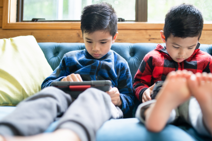 two asian children on their phones and ipads, sitting on a couch