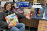 a librarian smiling and holding some books. She is gesturing to a table with more books on display in a library.