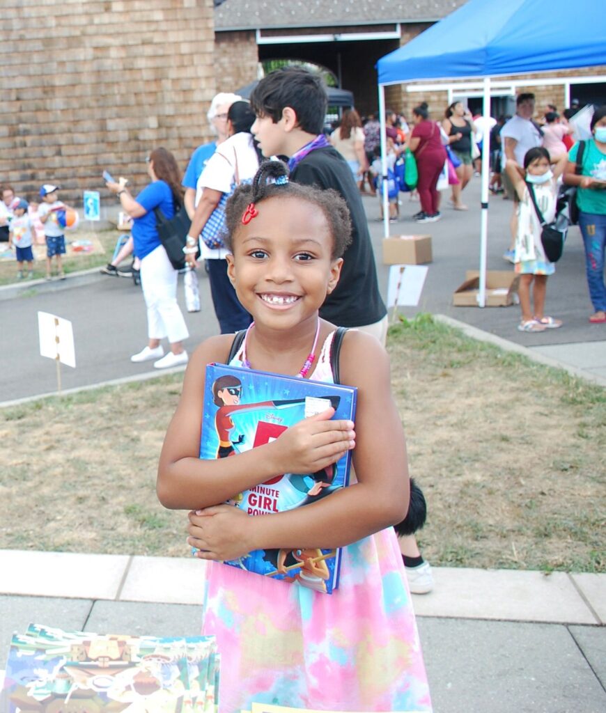 photo of a young girl hugging a book and smiling. Photo was taken outside at a book distribution event.