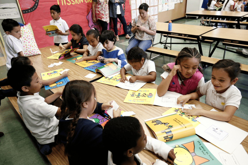 group of kids sitting at a long table, reading books