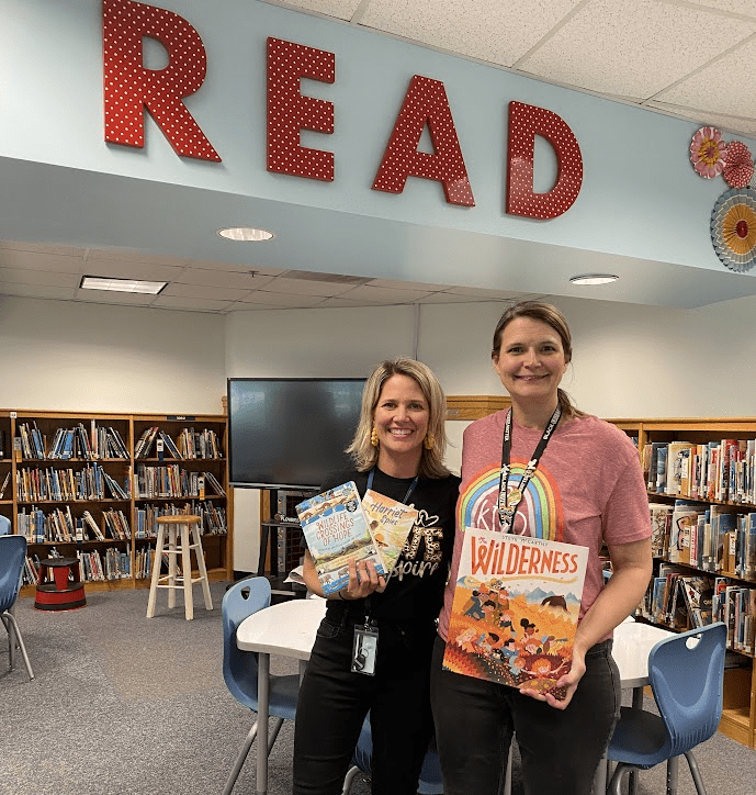 image of a two librarians standing in a school library, holding up books and smiling. Above them is a big sign that says, 'Read'. 