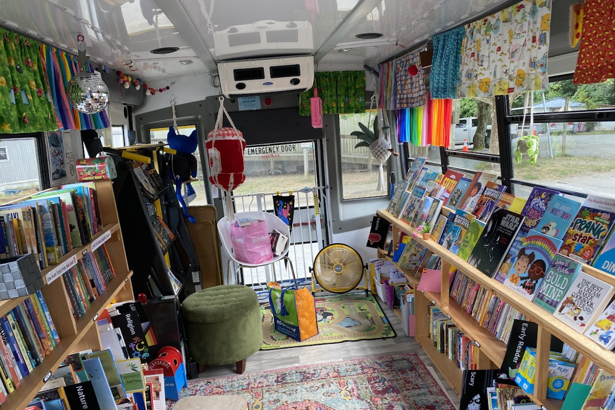 image of a converted school bus that now holds rows of books on each side. There is colorful decorations and the image is from the back of the bus, facing forward.