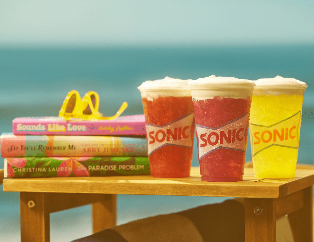 Three new Sonic drinks and three books by authors Ashley Poston, Abby Jimenez, and Christina Lauren on a wooden table at the beach.
