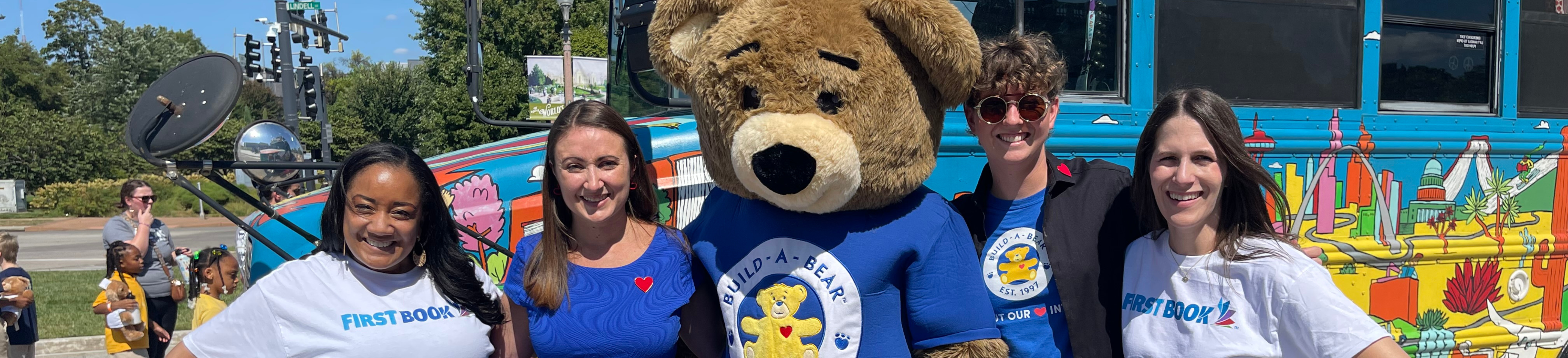 image of a group of people posing for a photo together in front of a colorful bus. In the middle of the group, is a mascot of Build-A-Bear.