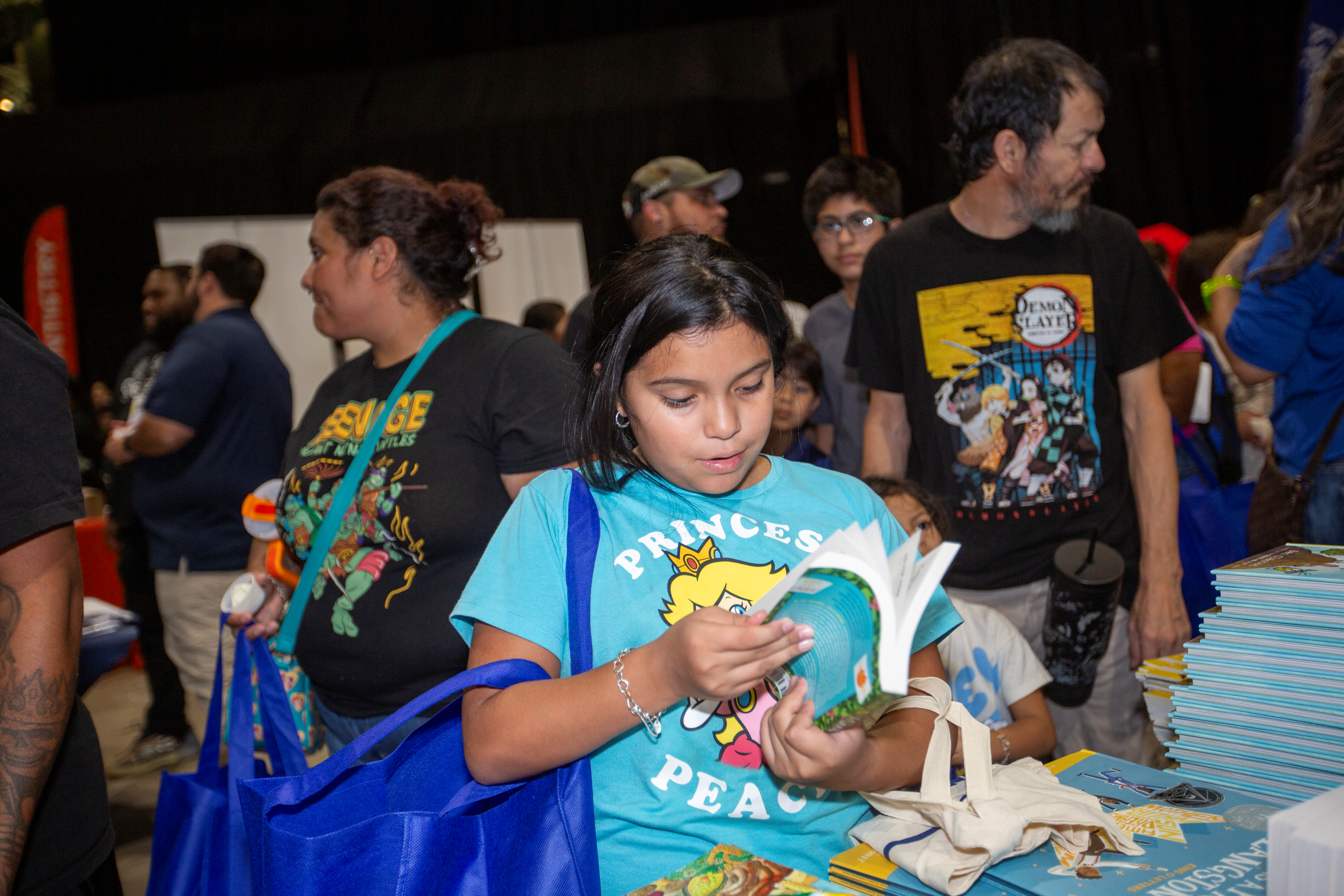 Young girl in a Princess Peach shirt reading a book at a community event, surrounded by people and stacks of books.
