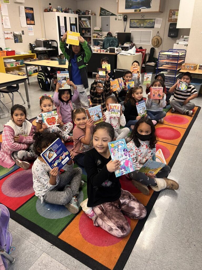 Young students sitting on a colorful classroom rug, smiling and holding up books they’ve received.