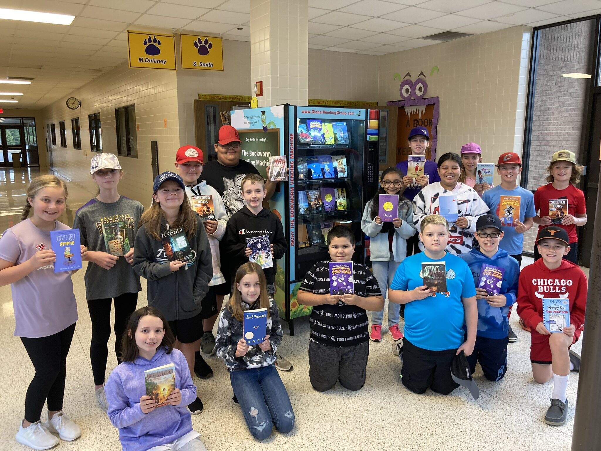 Group of older elementary students standing and kneeling in front of a book vending machine, proudly holding up their selected books in a school hallway.